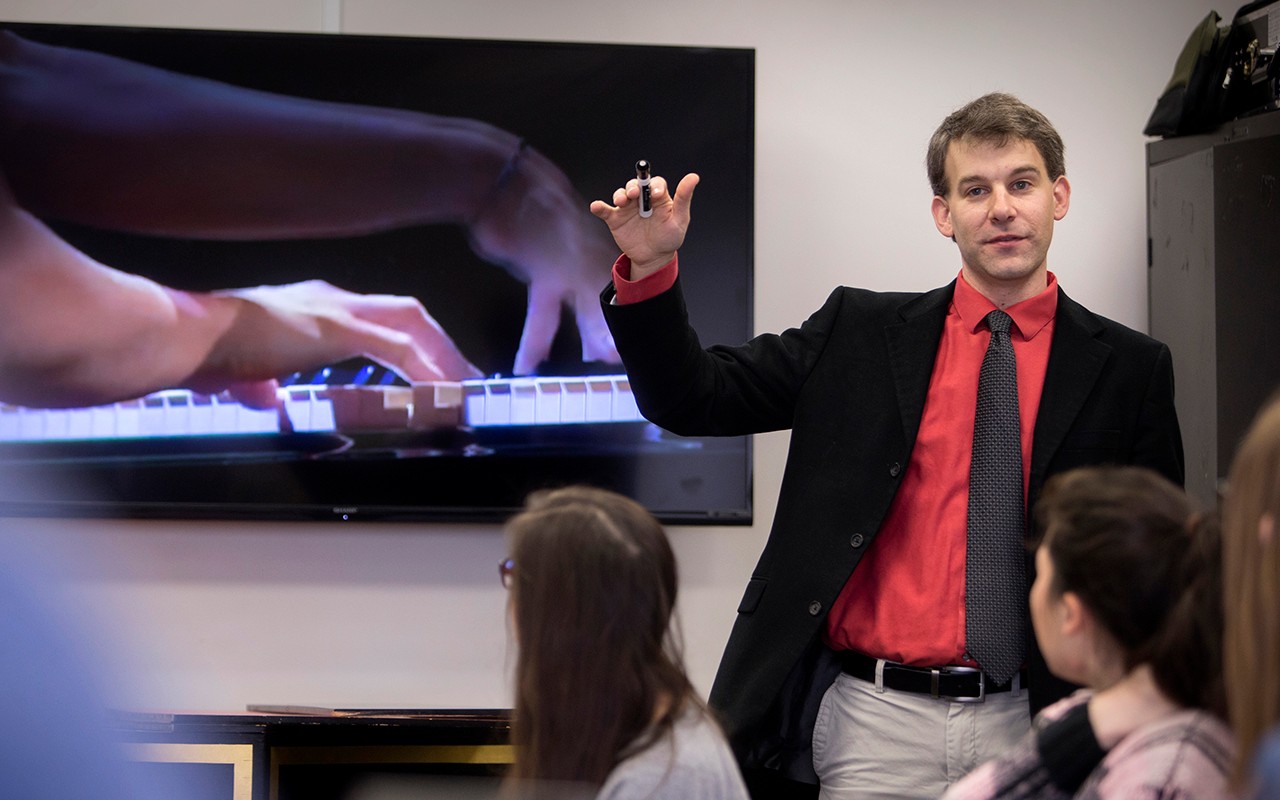 Musicology faculty member teaches a class in front of a video screen showcasing a pianist