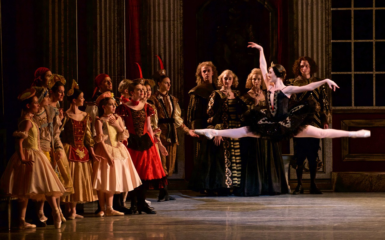 Ballerina leaps in a ballroom full of elaborately dressed spectators in a production of Swan Lake