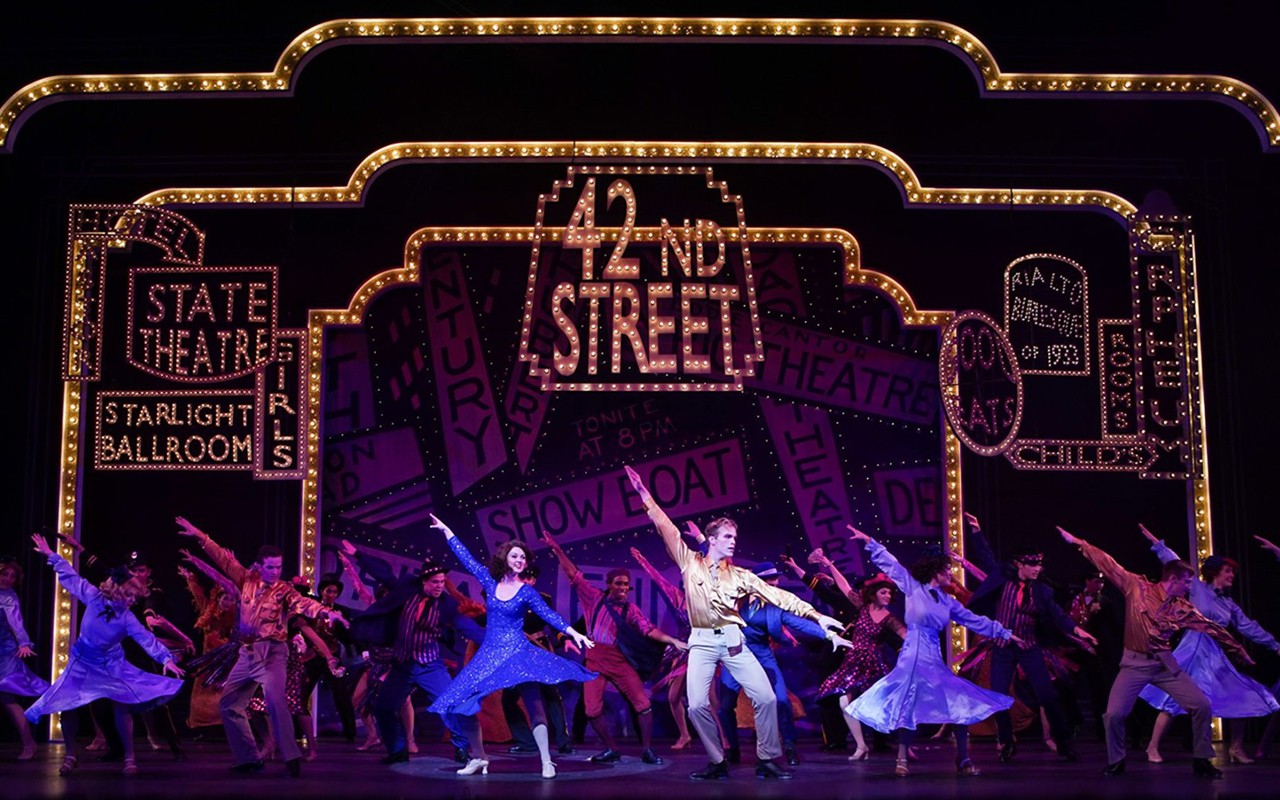 Large cast of student performers dance under lighted Broadway signs in a production of 42nd Street